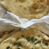 A picture of two pieces of naan. The one on the top is plain, with bubbles and brown spots. The one on the bottom has small pieces of minced garlic and chopped cilantro dotting the top, along with bubbles and brown spots.