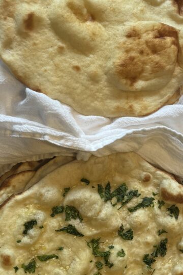 A picture of two pieces of naan. The one on the top is plain, with bubbles and brown spots. The one on the bottom has small pieces of minced garlic and chopped cilantro dotting the top, along with bubbles and brown spots.