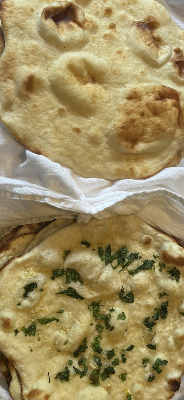 A picture of two pieces of naan. The one on the top is plain, with bubbles and brown spots. The one on the bottom has small pieces of minced garlic and chopped cilantro dotting the top, along with bubbles and brown spots.